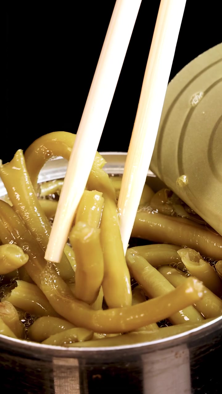 Chopsticks grasp and lift green beans from an open can under bright, direct lighting. The close-up shot emphasizes texture and movement against a black background
