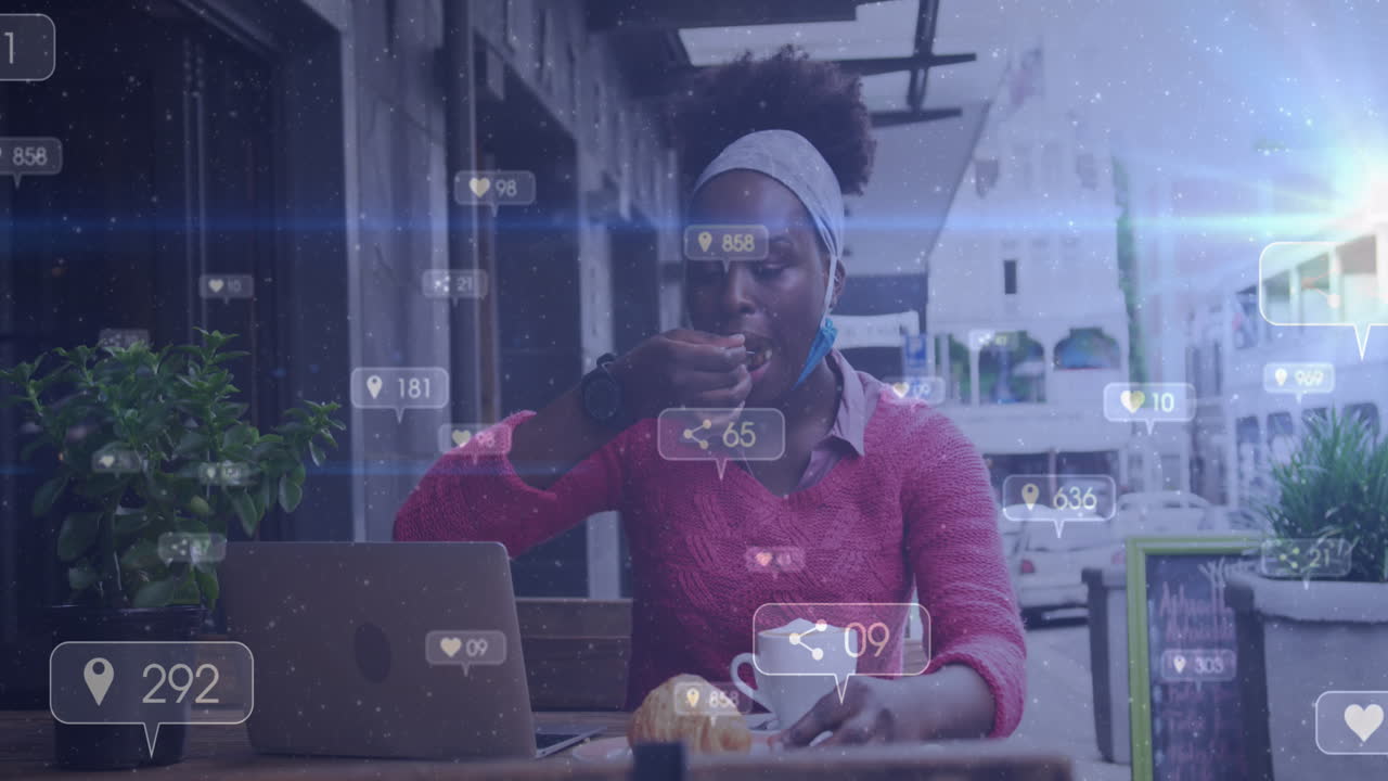 Animazione di notifiche di social media su donna afroamericana che usa il portatile e mangia in una caffetteria.