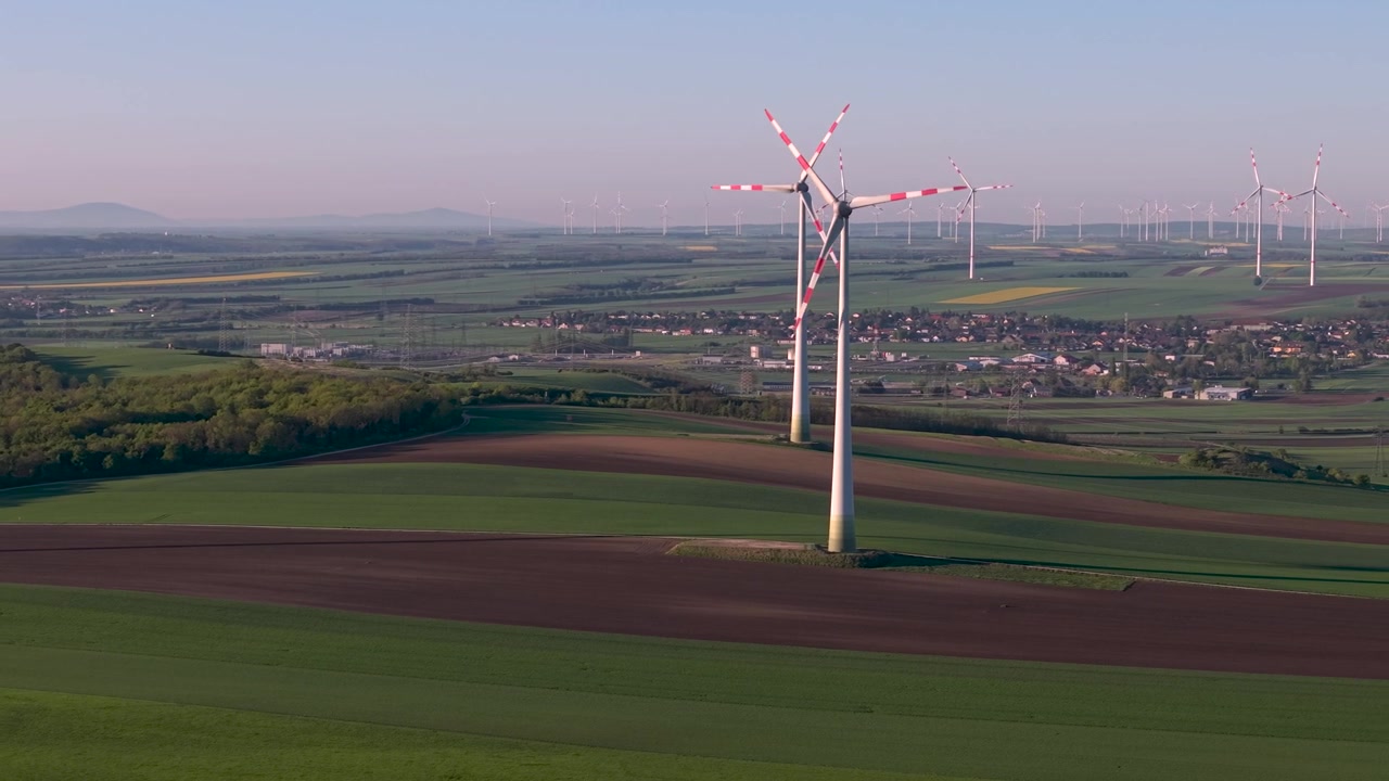 Vista aerea di un grande parco eolico che si estende su un paesaggio agricolo pianeggiante. Molteplici pale eoliche bianche con marcature rosse visibili attraverso campi verdi e una città lontana. Installazione di energia rinnovabile.