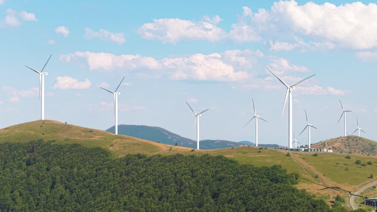 Beautiful drone footage of green energy turbines rotating on mountain slopes with blue sky and drifting clouds. Clean energy in scenic nature