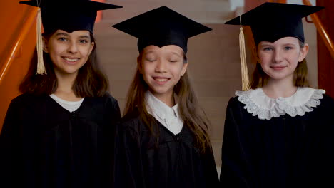 portrait of three happy preschool female students in cap and gown posing and looking at the camera at the graduation ceremony