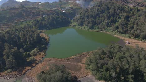 Aerial-wide-shot-of-Telaga-Warna-Lake-in-Dieng-Plateau-with-terraced-plantation-fields-on-hill-in-sunlight,-Indonesia