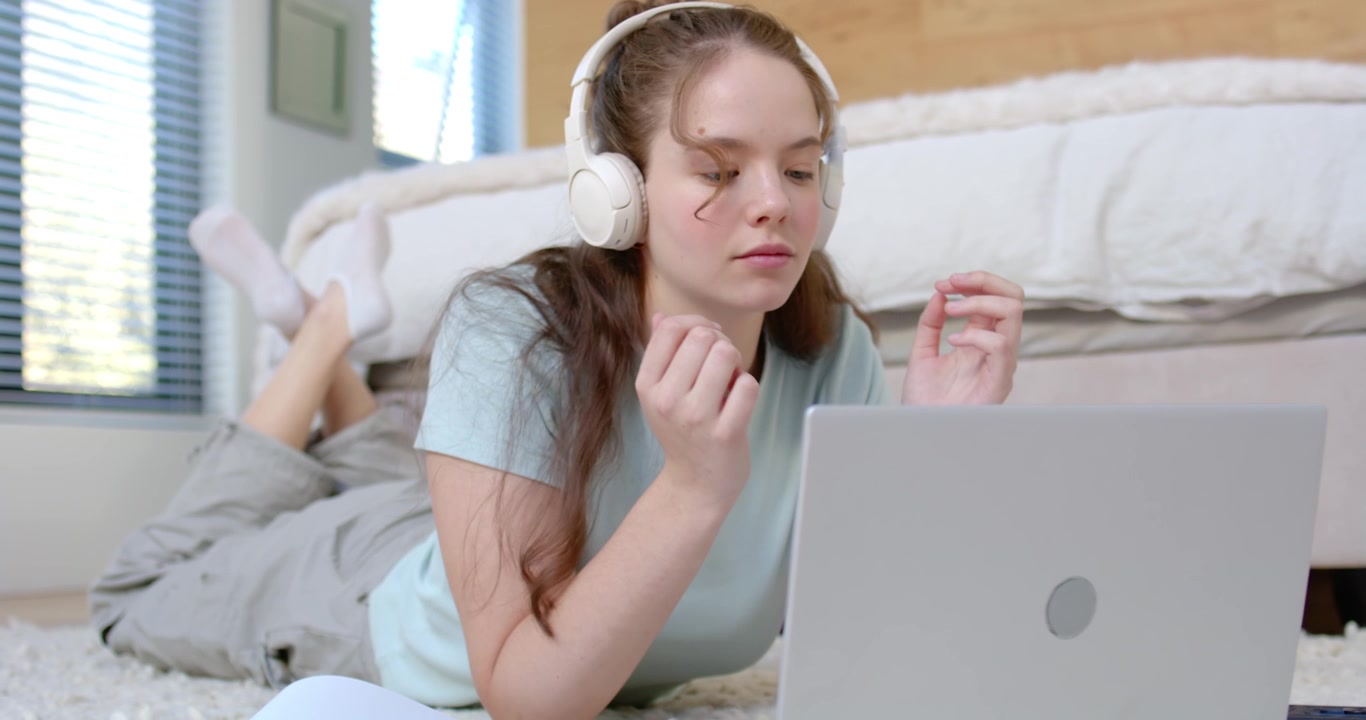 Donna che regola le cuffie con l'audio del laptop in riproduzione, chinandosi per digitare per lavoro su un tappeto in camera da letto.