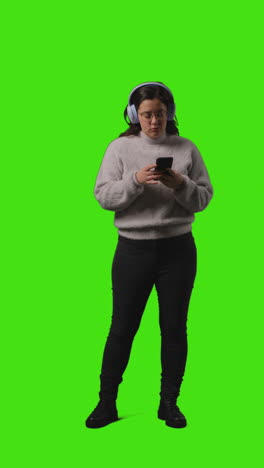 vertical video full length shot of woman wearing wireless headphones streaming music from mobile phone against green screen 2