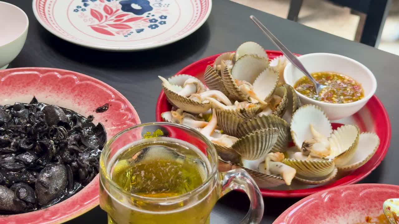 Steamed blood cockles, dipping sauce, and a glass of beer arranged on a table with patterned plates in bright natural daylight, static overhead view