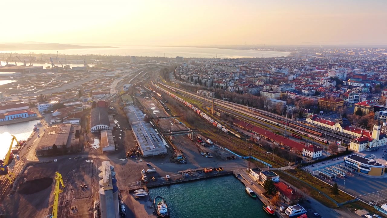 Solnedgångsljus över en hamnjärnvägsstation och stad, Burgas, Bulgarien. Varm gyllene kvällsdrönarvy över ett bangård, järnvägsspår, passagerarterminalbyggnad och stadsmiljö.