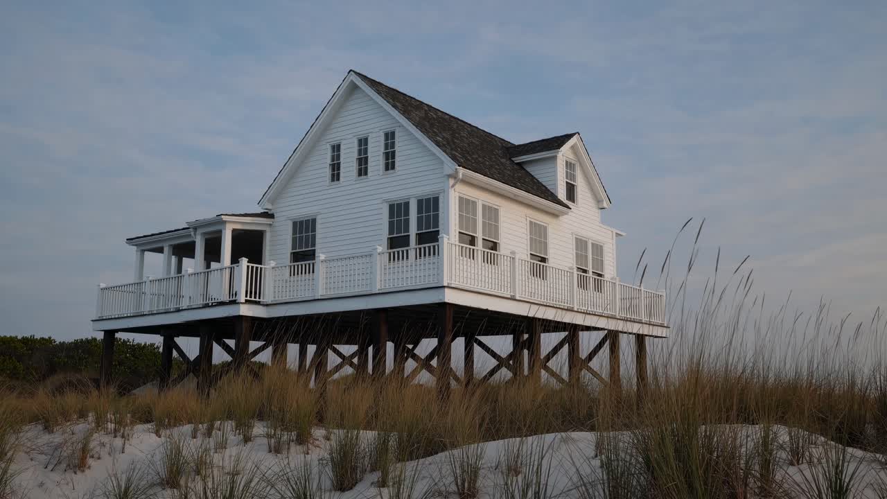 Weathered white beach house perched on wooden stilts, overlooking sandy dunes and coastal grasses during golden sunset lighting, embodying serene seaside architectural charm
