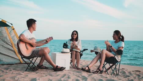 group of happy asian friends playing guitar and singing with clapping while picnic and camping on beach in outdoors vacation summer.