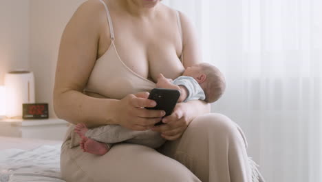 young mother breastfeeding her newborn baby and using mobile phone while sitting on the bed at home