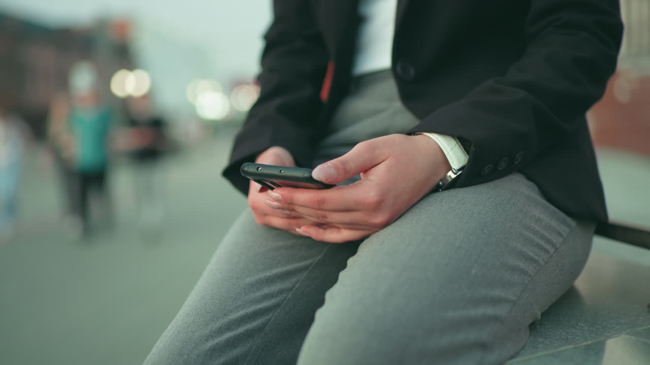 Close up partial view of lady in black suit seated with phone in hand, grey trousers, white watch, and blur bokeh background featuring people walking on street in soft light urban setting