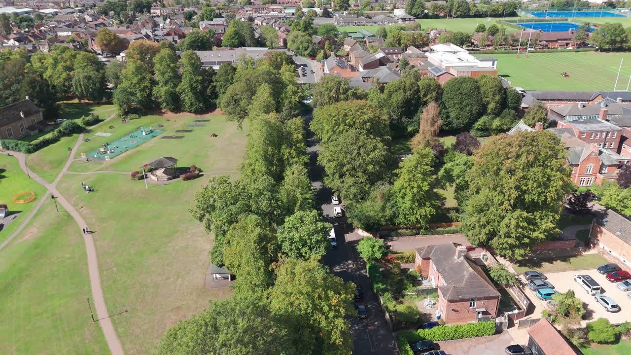 An aerial shot of a park, recreation area, next to a tree-lined, shaded street. In the background are residential housing and what appears to be a school, and a sports field