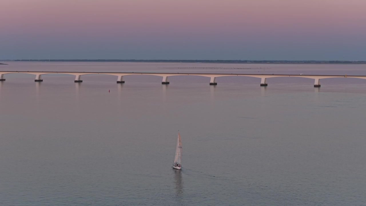 Sejlbåd, der sejler på roligt vand under en bro ved solnedgang.