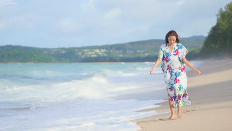 Hermosa-Joven-Modelo-Asiática-Caminando-Descalza-En-La-Playa-De-La-Isla-De-Arena-Blanca-Usando-Un-Vestido-De-Verano-Cuando-Las-Olas-Espumosas-Se-Estrellan,-En-Cámara-Lenta-Durante-El-Día