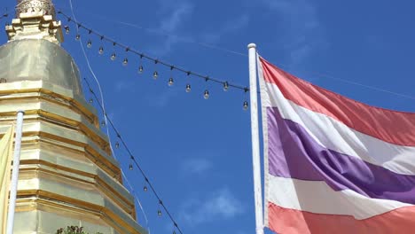 a thai flag flutters next to a golden stupa under a clear blue sky.
