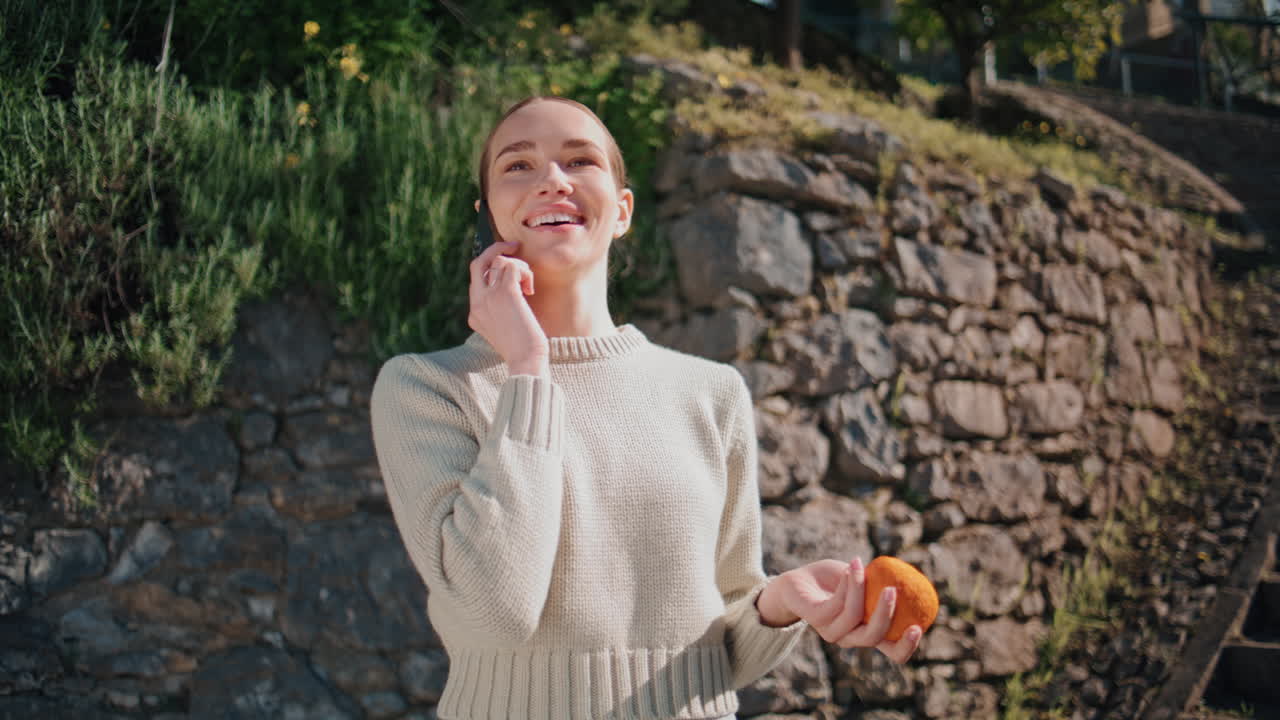 Calling woman sniffing orange at sunny park closeup. Carefree lady talking phone
