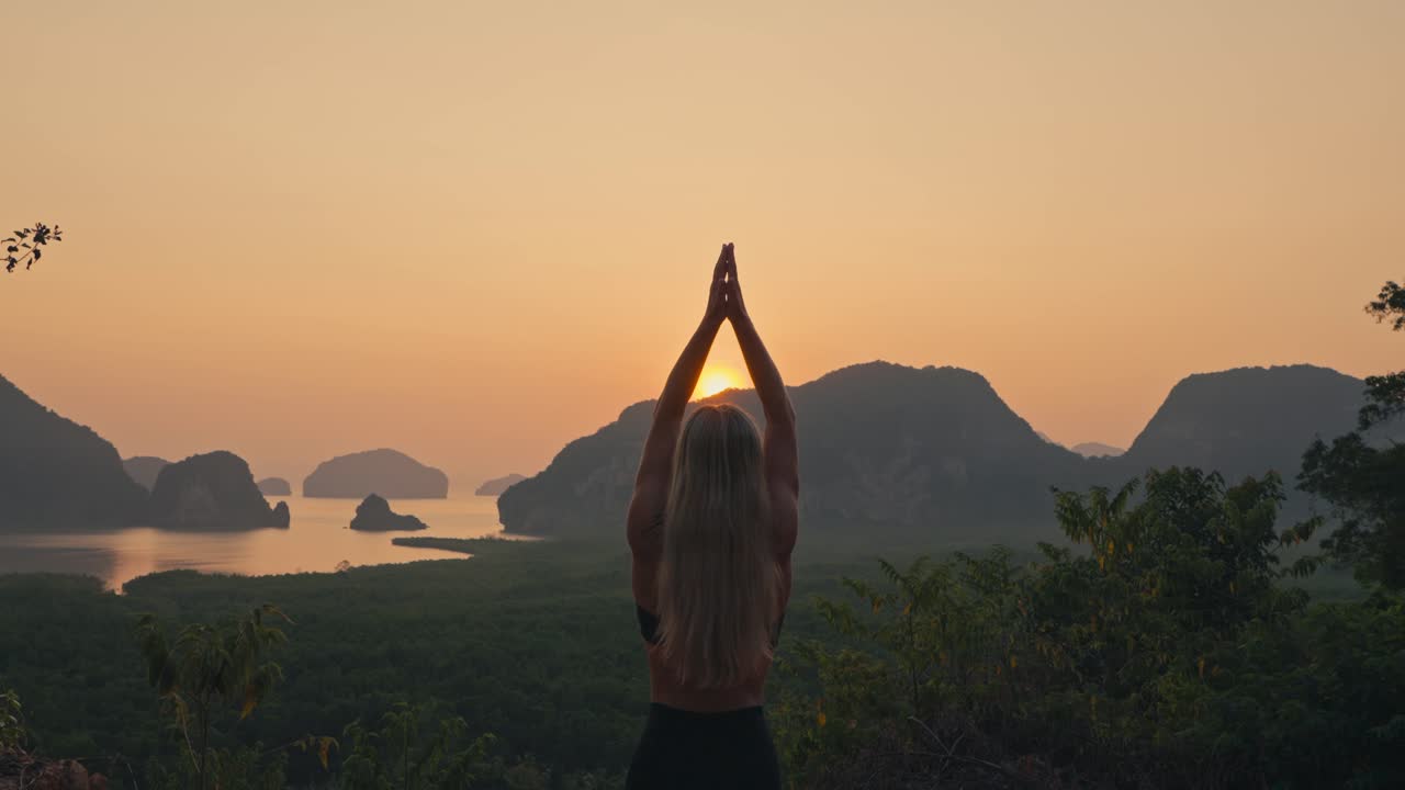 Piano medio di una donna che esegue la posizione yoga di preghiera in saluto verso l'alto, affacciata su una baia con isole tropicali, con un bagliore dorato nel cielo e il sole che spunta dietro le montagne.