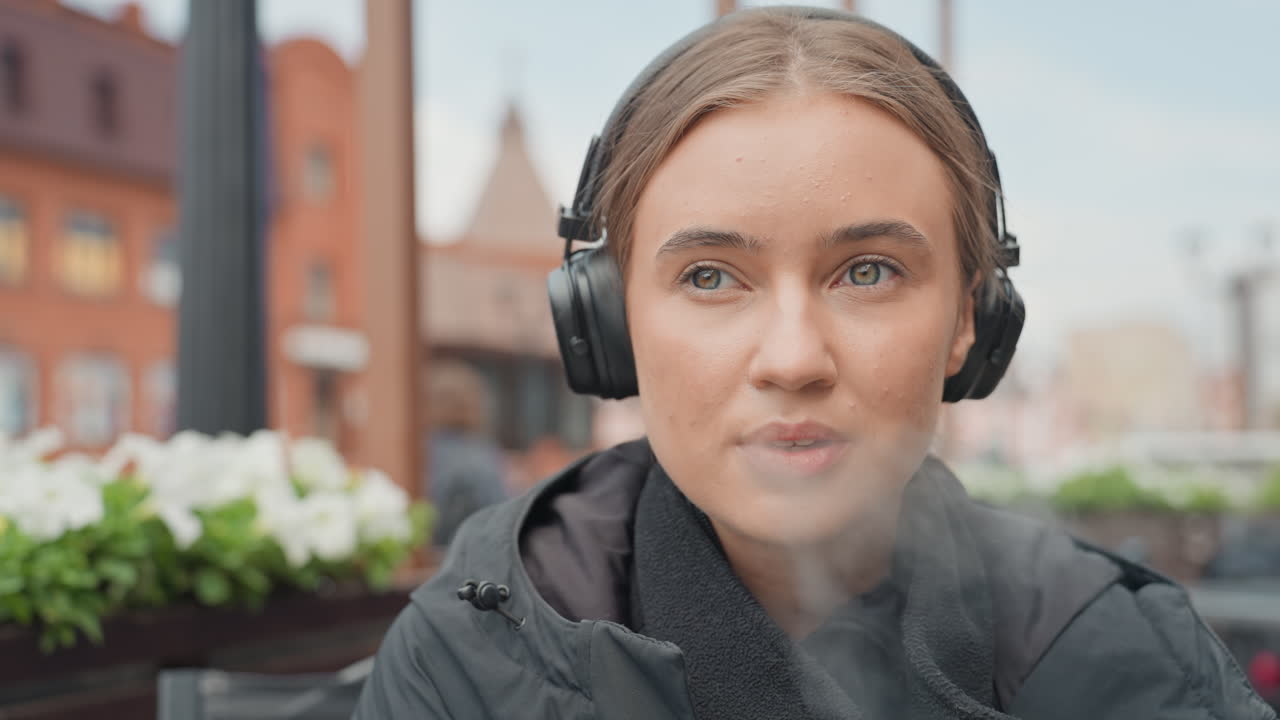 Ung kvinne med hodetelefoner utendørs som sitter på en kaféterrasse med murbygninger og blomsterkasser, tar et drag fra en vape, skifter uttrykk fra oppmerksom til avslappet, øynene lukkes i et rolig øyeblikk