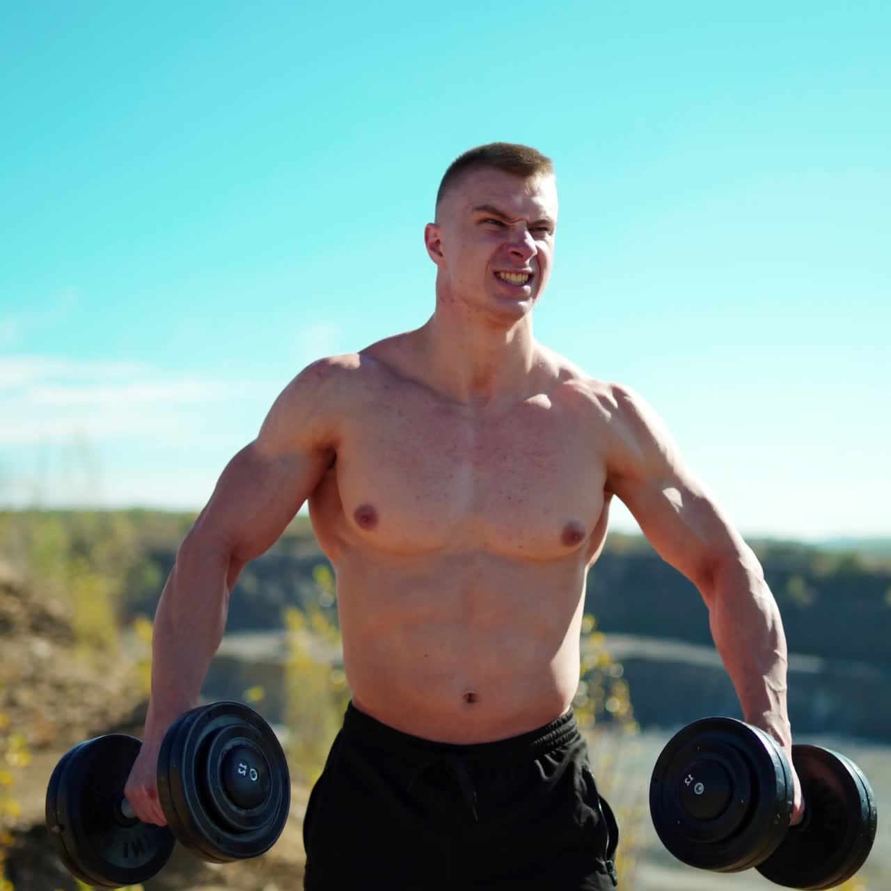 Portrait of concentrated sportsman lifts dumbbells. Strong bodybuilder training with heavy dumbbells on the nature background in summer.