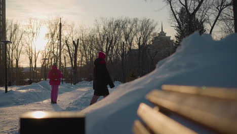 snowy path with golden sunlight reflecting on bench as two people in winter clothing walk past each other in opposite directions, surrounded by snow piles, street lamps, and leafless trees at sunset