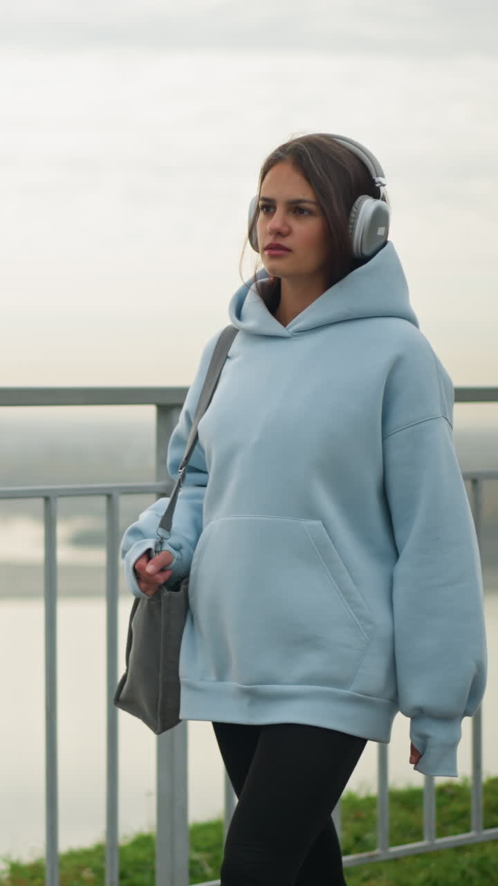 Young girl in blue hoodie with headphones walking beside river with iron rail demarcating her and water, scenic peaceful outdoor setting with flowing river and serene background view