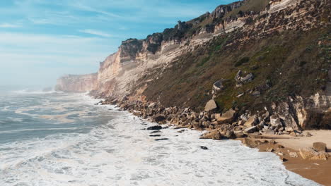 Toma-Aérea-Hacia-Atrás-De-Olas-Espumosas-Que-Llegan-A-La-Playa-De-Arena-De-Nazare-Con-Una-Gigantesca-Formación-De-Acantilados-En-El-Fondo