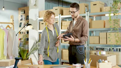 caucasian young designer man typing on tablet and speaking with female coworker about new clothing collection in clothing shop warehouse