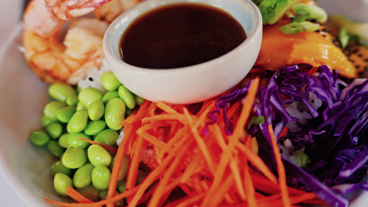 Close up of a poke bowl with rice, avocado, carrot, red cabbage, shrimp and mango and sauce in a white bowl