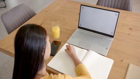 mujer biracial asiática en la mesa escribe, con portátil y teléfono inteligente cerca, espacio de copia