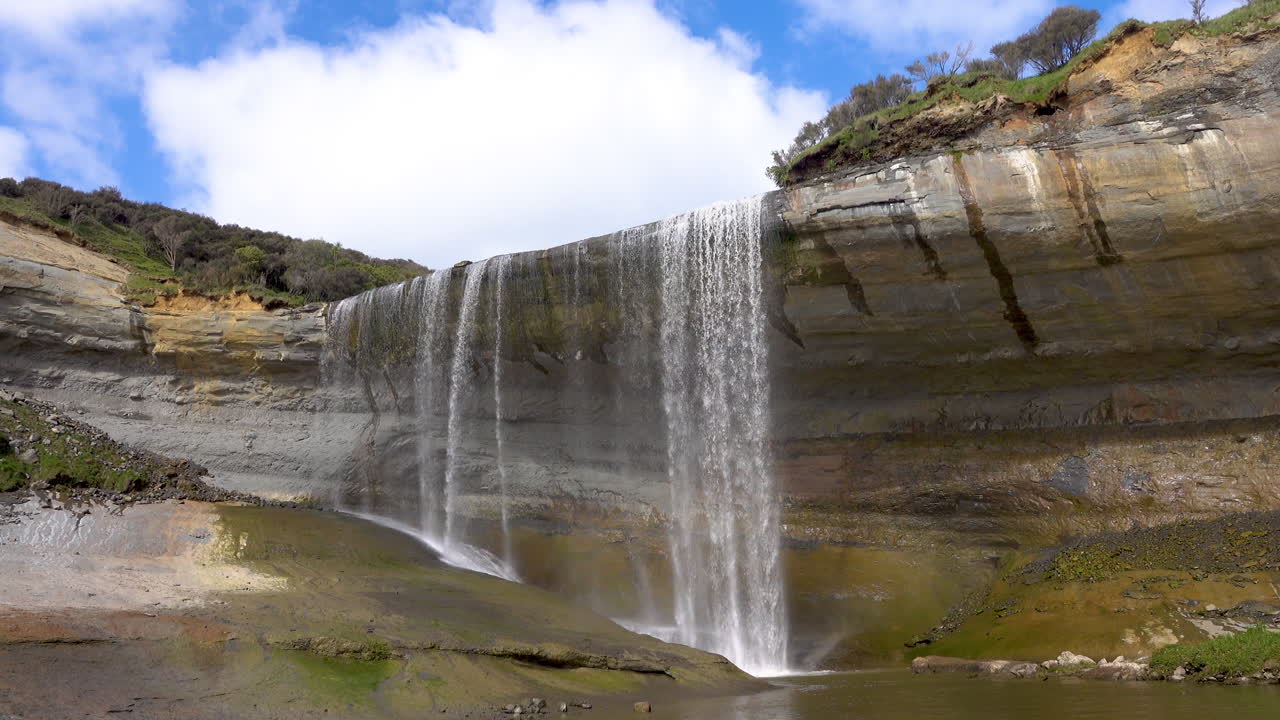 toma estática de cascadas de mangatiti que fluyen desde un acantilado rocoso contra el cielo azul y las nubes