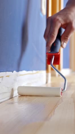 vertical view of a person applying varnish with a roller along a wooden floor and taped baseboard, part of an interior renovation project with precise detailing and clean finishing