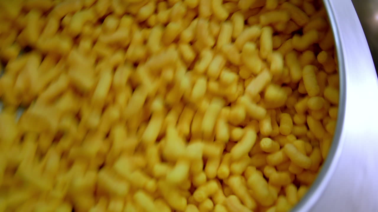Plenty of sweet corn snacks being mixed in metal tank. Close up. Manufacturing maize crispy sticks at factory.