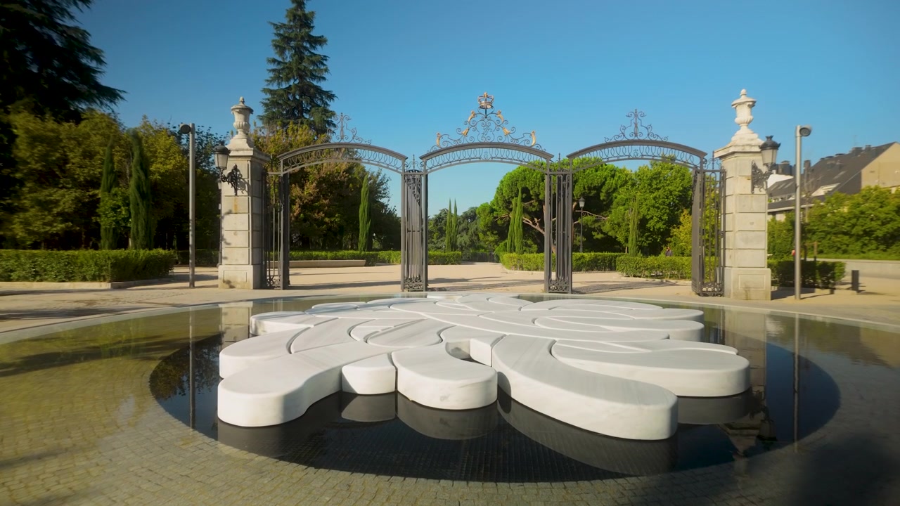 A minimalist white stone sculpture sits in a shallow reflecting pool at the entrance of a lush public park in Madrid. Suitable for travel and urban design content.