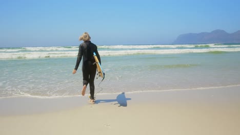 Vista-Trasera-De-Una-Surfista-Caucásica-Senior-Activa-Corriendo-En-El-Mar-Bajo-El-Sol-4k