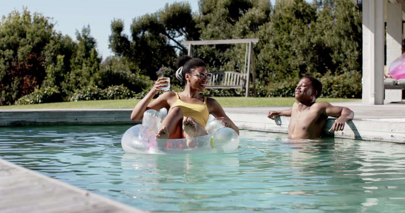 African American couple smiling, woman in yellow swimsuit raising drink in clear ring at home pool