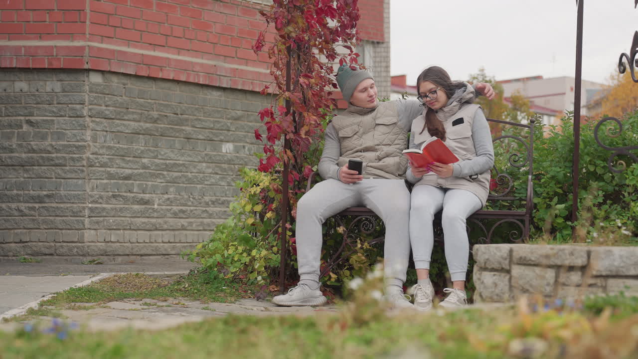 Two couple seated outdoors in cool breeze as man warmly brings woman closer, she reads book while he holds phone, surrounded by greenery, flowers, and brick wall in peaceful urban park setting
