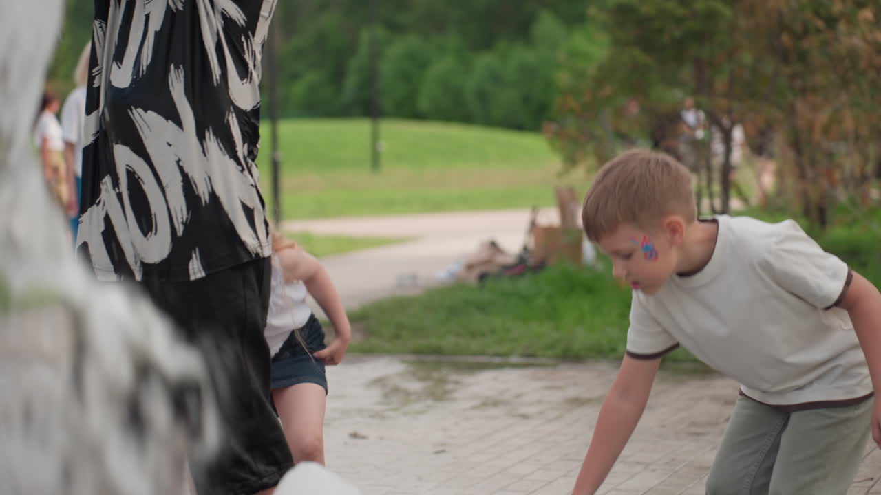 Outdoor children near water fountain bending to pick object from wet pavement, blurred adults and other kids in busy green park, summer day activity showing curiosity and interaction