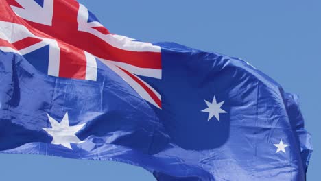 Australian national flag ripples dynamically in bright daylight, captured in close-up with clear blue sky background and smooth camera tracking movement