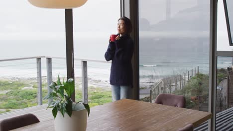 Happy-asian-woman-drinking-coffee-and-thinking-at-window-alone