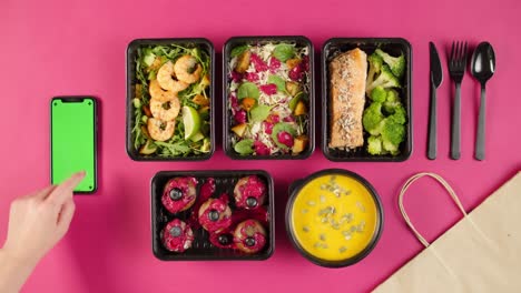 food delivery top view, take away meals in disposable containers on pink background. lunch boxes with cooked vegetarian dishes, using phone with chroma green screen. healthy diet. catering service