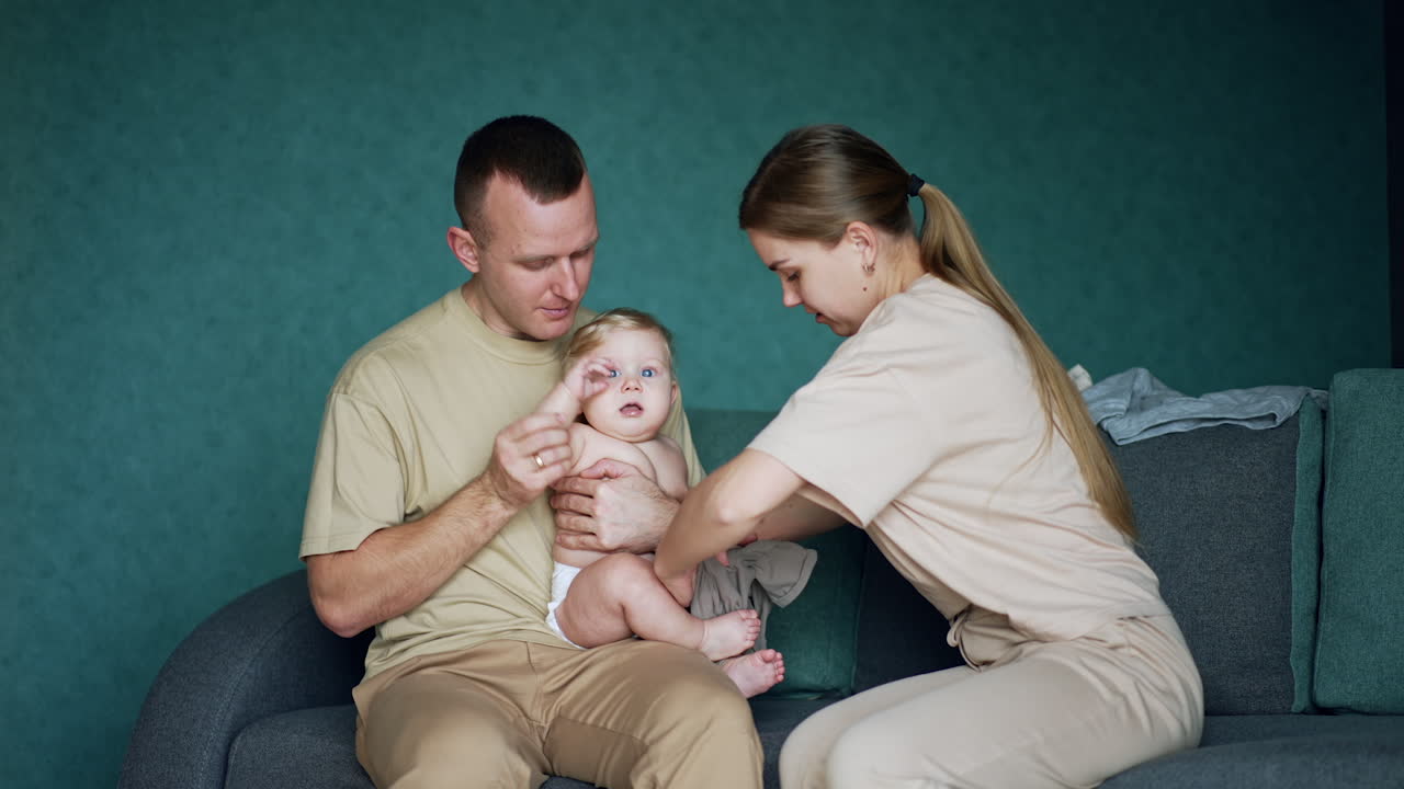 Loving dad holding a cute blond bare child. Long-haired woman puts on pants on her baby boy. Blue backdrop.