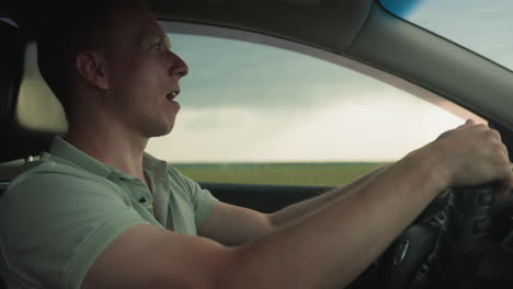 side view man driving car looks out window at vast green field under dramatic cloudy sky while hands grip wheel in quiet interior shot capturing serene travel moment in countryside road trip