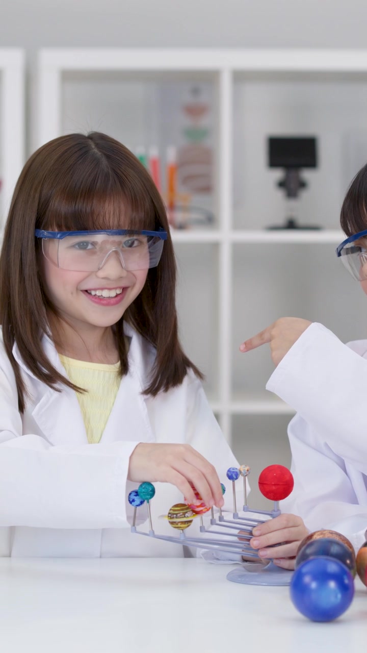 Young students wearing safety goggles and lab coats examine a solar system model in school.
