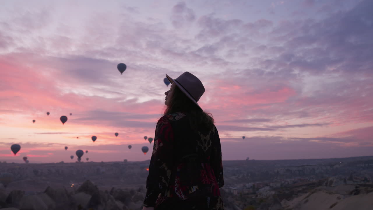 Beautiful Female Tourist In Dress And Hat Watching Hot Air Balloons Flying In The Sky At Sunrise Over Cappadocia In Turkey