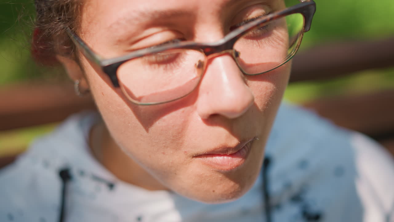 벤치에 앉아 허브 맛을 보는 여성의 클로즈업, 맛을 테스트하는 홈 셰프, 연구를 위해 시식하는 학생, 햇빛이 비치는 공원에서 천천히 음미하는 사려 깊은 사람, 안경에 비친 모습, 후드티 디테일, 부드럽게 씹는 모습, 잎의 질감