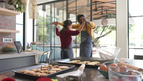 Feliz-Pareja-Diversa-En-Delantales-Horneando-Galletas-Y-Bailando-En-Una-Cocina-Soleada,-Copiando-Espacio,-Cámara-Lenta