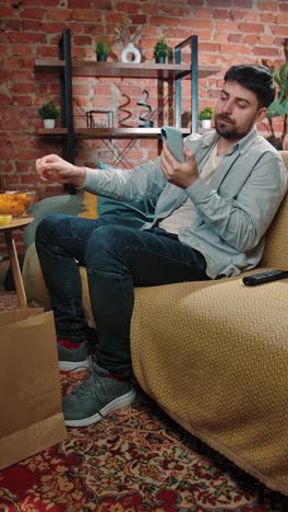 man relaxing on a couch, eating snacks and watching a smartphone