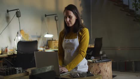 caucasian female jeweller in workshop wearing apron, sitting at desk, using laptop