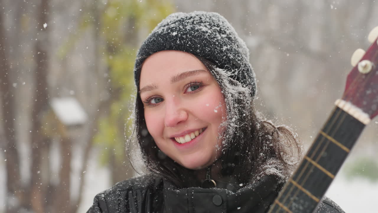 눈 인 공원에서 카메라를 향해 따뜻하게 미소 짓는 젊은 여성, 활기찬 빨간색 어쿠스틱 기타를 들고, 검은색 베니와 재에 눈을 고, 얼어붙은 나무로 둘러싸여 있습니다.