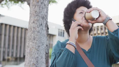 mujer biracial feliz de talla plus hablando en el teléfono inteligente y bebiendo café