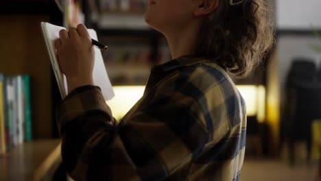 Confident-girl-with-curly-hair-wearing-glasses-takes-inventory-of-books-on-shelves-in-library